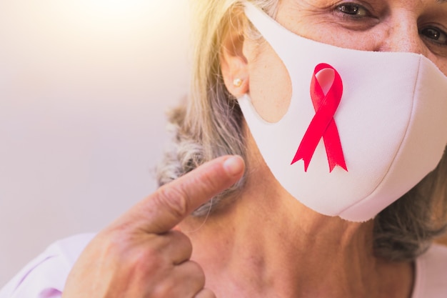 Femme mature avec masque pandémique et ruban rose isolé sur blanc. image du mois de la sensibilisation au cancer du sein.