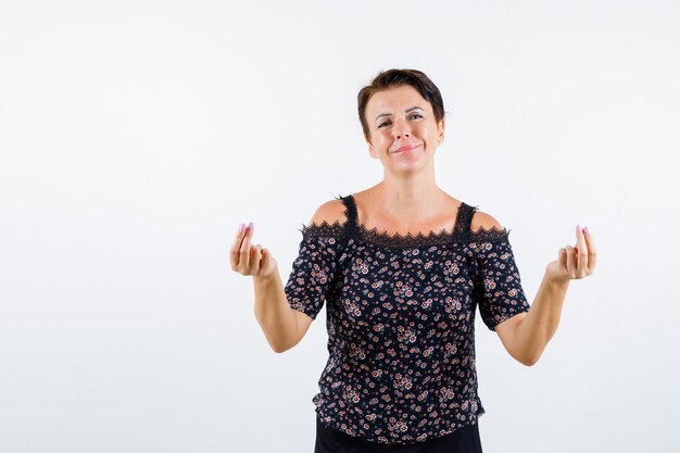 Femme mature montrant le geste de l'argent en chemisier floral et jupe noire et à la joyeuse, vue de face.