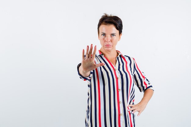 Femme mature montrant le geste d'arrêt, tenant la main sur la taille en chemisier rayé et à la grave, vue de face.