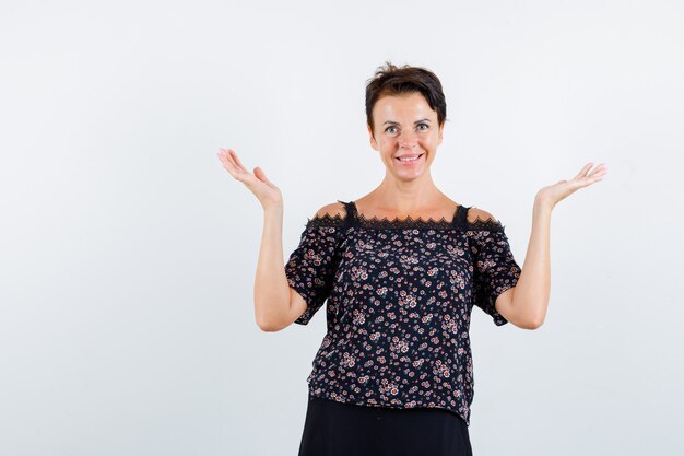 Femme mature montrant un geste impuissant en chemisier floral et jupe noire et à la vue de face, joyeuse.