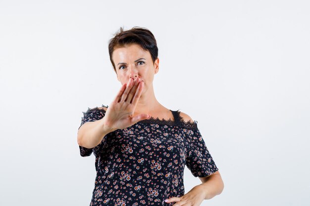 Femme mature montrant le panneau d'arrêt, tenant la main sur la taille en chemisier floral, jupe noire et à la confiance. vue de face.