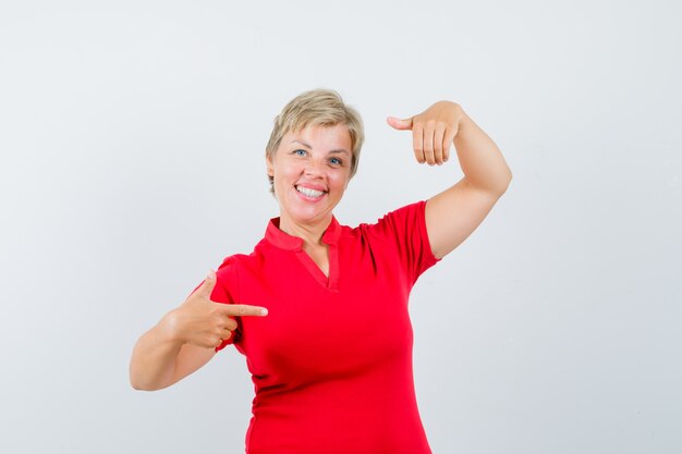 Femme mature pointant de côté, faisant semblant de tenir quelque chose en t-shirt rouge et à la confiance