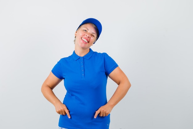 Femme mature pointant vers le bas, clignant des yeux en t-shirt bleu et à la bonne humeur