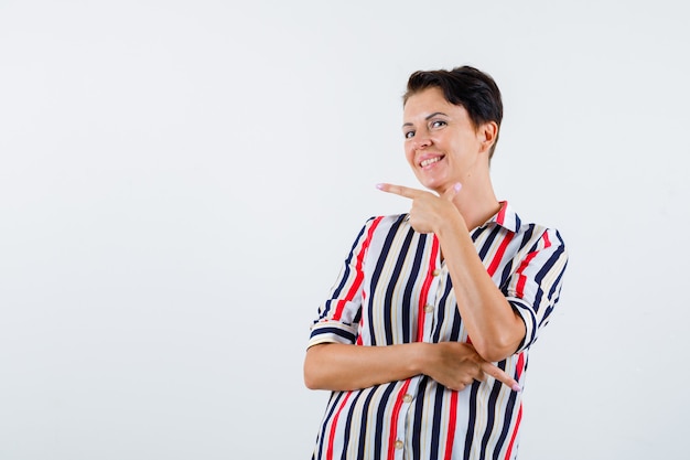 Femme mature pointant vers la gauche et la droite avec l'index en chemisier rayé et à la bonne humeur. vue de face.