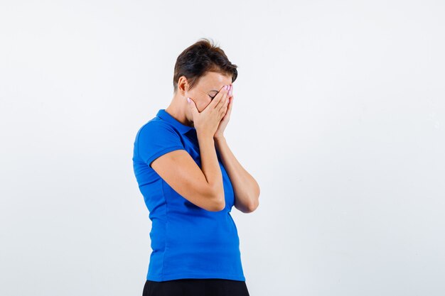 Femme mature en t-shirt bleu couvrant le visage avec les mains et regardant déprimé, vue de face.
