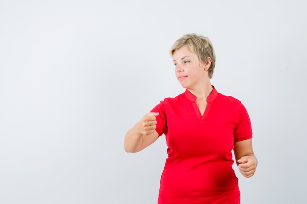 Femme mature en t-shirt rouge faisant semblant de tenir quelque chose, regardant de côté.