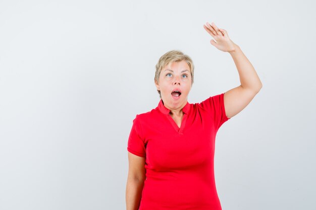 Femme mature en t-shirt rouge en levant la main pour montrer la hauteur et à la stupéfaction