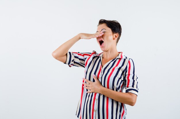 Femme mature tenant la main au-dessus de la bouche, essayant d'éternuer en chemisier rayé et à la vue épuisée, de face.