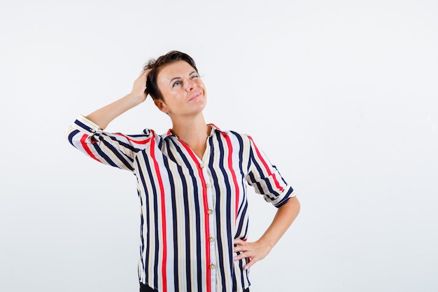 Femme mature tenant une main sur la taille, une autre main sur la tête, pensant à quelque chose en chemise rayée et à la pensif. vue de face.