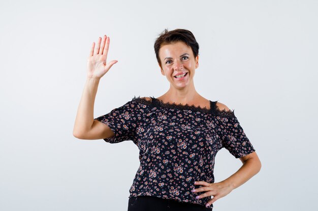 Femme mature tenant la main sur la taille, montrant le panneau d'arrêt en chemisier floral, jupe noire et à la joyeuse. vue de face.