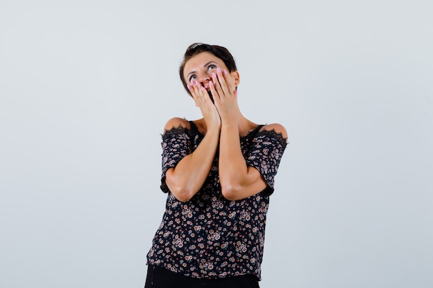 Femme mature tenant les mains sur les joues tout en regardant en chemisier et à la vue étonnée, de face.