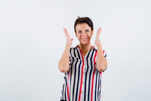 Femme mature tenant les mains près de la tête, souriant en chemisier rayé et à la flottabilité. vue de face.