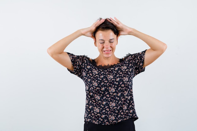 Femme mature tenant les mains sur la tête, regardant vers le bas en chemisier floral, jupe noire et à la jolly, vue de face.