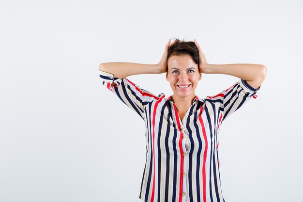 Femme mature tenant la tête avec les mains en chemise rayée et à la vue de face, heureux.