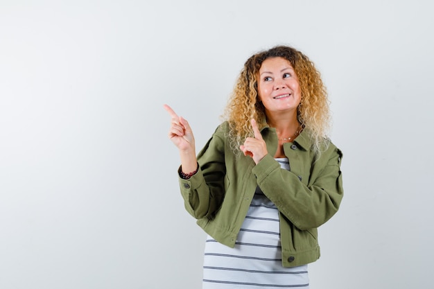 Femme mature en veste verte, t-shirt pointant vers le haut tout en souriant et à la joyeuse, vue de face.