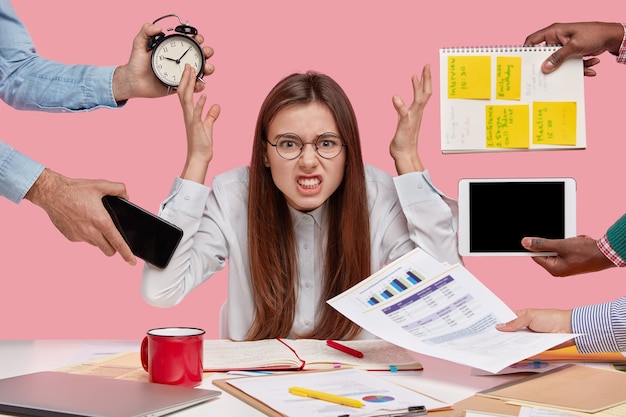 Photo gratuite une femme maussade se tient la main avec dégoût, étant submergée par beaucoup de travail, ressent la pression de ses collègues, s'assoit au bureau avec de la documentation