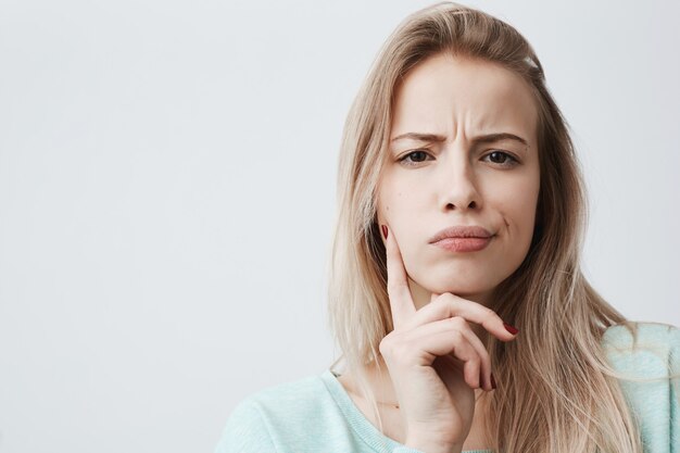 une femme mécontente aux cheveux blonds a une expression indignée du visage, fronce les sourcils, ne peut pas comprendre quelque chose. Attractive femme insatisfaite perplexe garde la main sur le menton