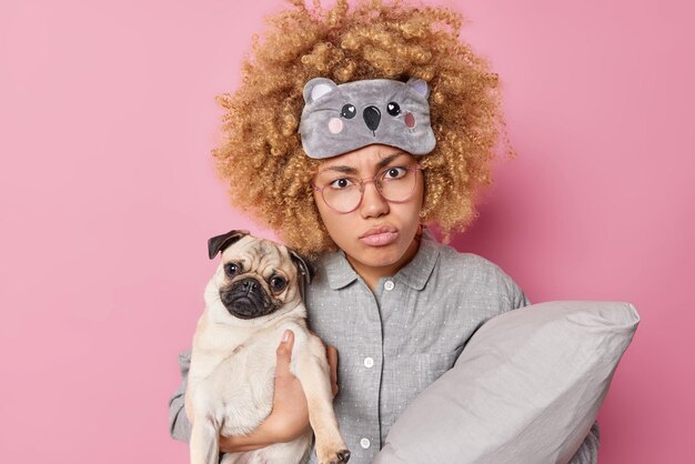 Une femme mécontente aux cheveux bouclés vêtue d'un pyjama et d'un masque de sommeil tient un oreiller se prépare pour le sommeil tient des poses de chien carlin sur fond rose aime la compagnie du meilleur ami Concept d'amitié