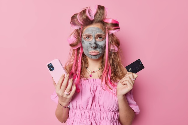 Une femme mécontente et frustrée n'a pas d'argent sur son compte bancaire détient une carte de crédit et un smartphone applique un masque de beauté sur le visage porte des bigoudis isolés sur fond rose utilise une application de shopping