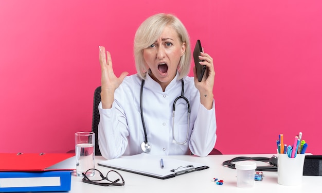 femme médecin adulte agacée en robe médicale avec stéthoscope assis au bureau avec des outils de bureau tenant un téléphone et criant sur quelqu'un isolé sur un mur rose avec espace de copie