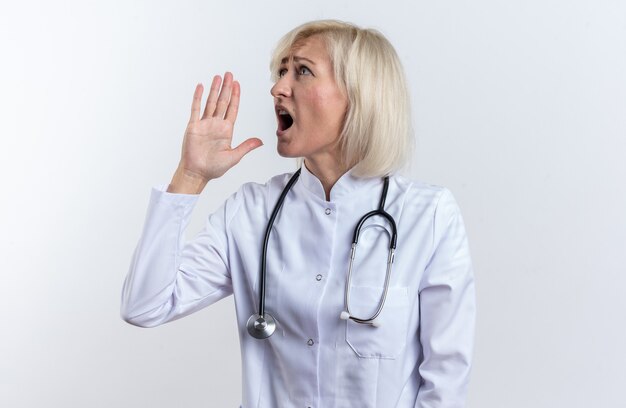 femme médecin adulte anxieuse en robe médicale avec stéthoscope tenant la main près de sa bouche regardant de côté appelant quelqu'un isolé sur un mur blanc avec espace de copie