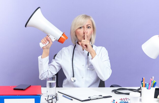 femme médecin adulte confiante en robe médicale avec stéthoscope assise au bureau avec des outils de bureau tenant un haut-parleur et faisant un geste de silence isolé sur un mur violet avec espace de copie