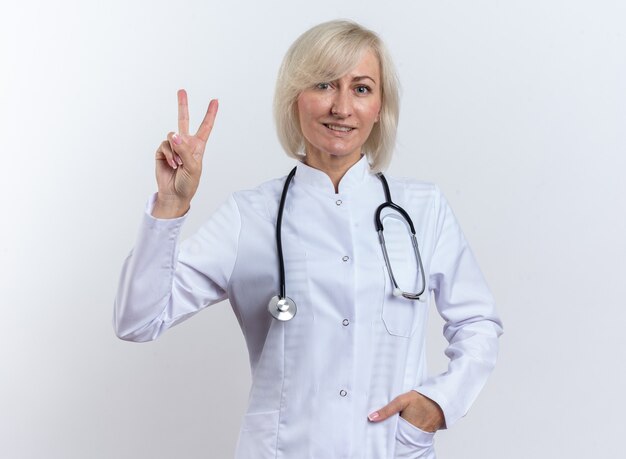 femme médecin adulte souriante en robe médicale avec stéthoscope gesticulant signe de victoire isolé sur mur blanc avec espace de copie