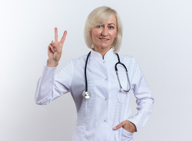 femme médecin adulte souriante en robe médicale avec stéthoscope gesticulant signe de victoire isolé sur mur blanc avec espace de copie