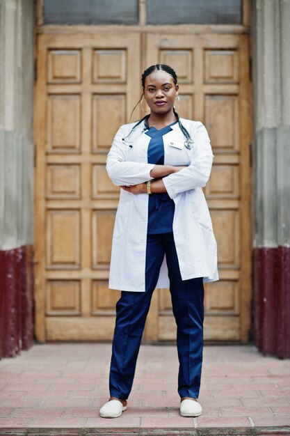 Femme médecin afro-américaine en blouse de laboratoire avec stéthoscope en plein air contre la porte de la clinique