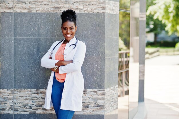 Femme médecin afro-américaine en blouse de laboratoire avec stéthoscope posé en plein air contre la clinique