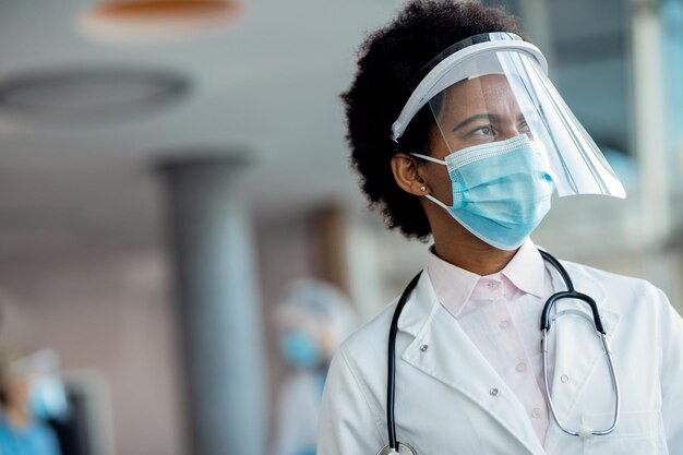 Femme médecin afro-américaine pensive avec écran facial et masque à l'hôpital
