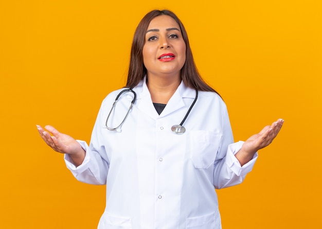 Femme médecin d'âge moyen en blouse blanche avec stéthoscope confus écartant les bras sur les côtés à la recherche