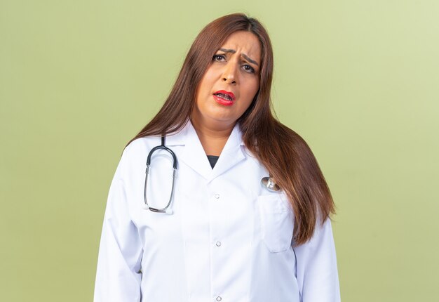 Femme médecin d'âge moyen en blouse blanche avec stéthoscope confuse et très anxieuse debout sur le vert