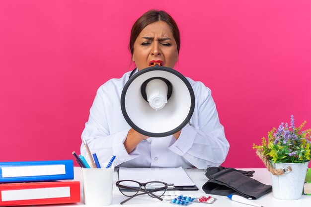 Femme médecin d'âge moyen en blouse blanche avec stéthoscope criant au mégaphone étant excitée assise à la table avec des dossiers de bureau sur rose