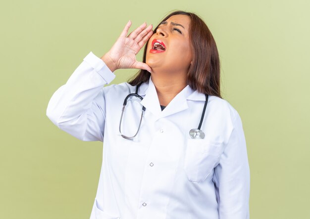Femme médecin d'âge moyen en blouse blanche avec stéthoscope criant fort avec la main près de la bouche debout sur un mur vert