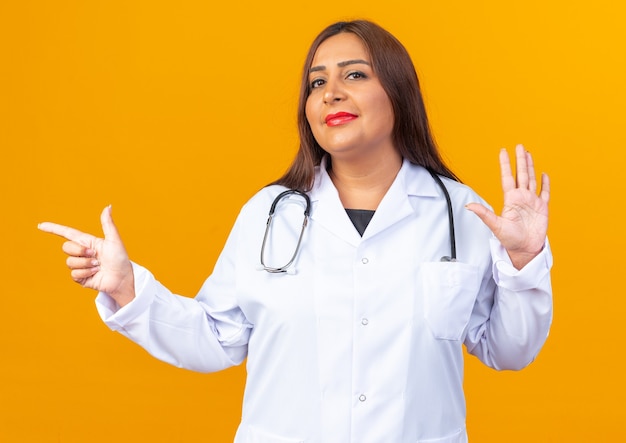 Femme Médecin D'âge Moyen En Blouse Blanche Avec Stéthoscope Heureux Et Positif Montrant Le Numéro Cinq Et Pointant Avec L'index Sur Le Côté Debout Sur Le Mur Orange