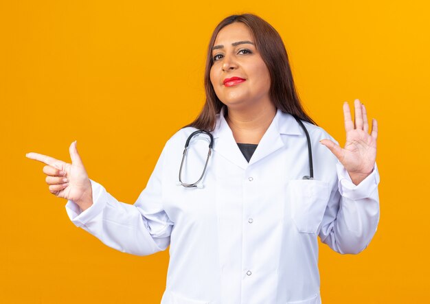 Femme médecin d'âge moyen en blouse blanche avec stéthoscope heureux et positif montrant le numéro cinq et pointant avec l'index sur le côté debout sur le mur orange