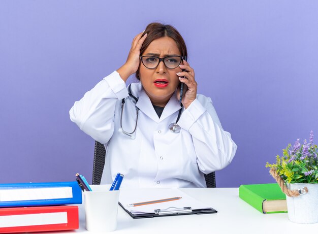 Femme médecin d'âge moyen en blouse blanche avec stéthoscope portant des lunettes semblant confuse tout en parlant au téléphone portable avec la main sur la tête pour erreur assise à la table sur le mur bleu