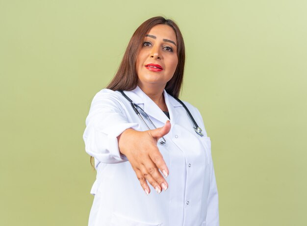 Femme médecin d'âge moyen en blouse blanche avec stéthoscope regardant l'avant souriant confiant offrant la main faisant un geste de salutation debout sur un mur vert