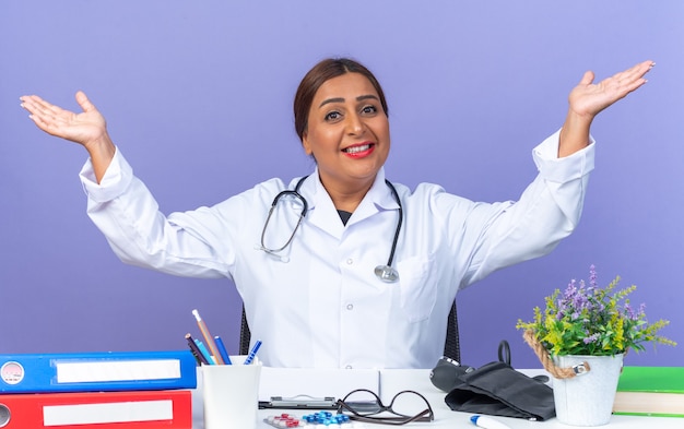 Femme médecin d'âge moyen en blouse blanche avec stéthoscope regardant à l'avant souriant heureux et positif écartant joyeusement les bras sur les côtés assis à la table sur le mur bleu