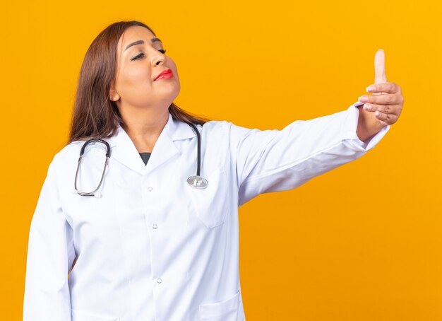 Femme médecin d'âge moyen en blouse blanche avec stéthoscope regardant de côté avec un sourire confiant sur le visage montrant les pouces vers le haut