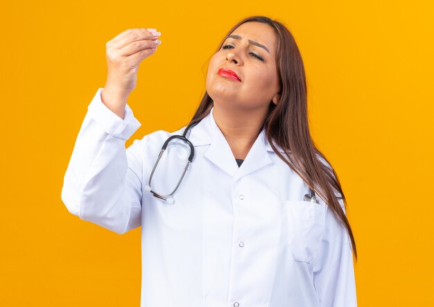 Femme médecin d'âge moyen en blouse blanche avec stéthoscope regardant ses doigts faire un geste d'argent