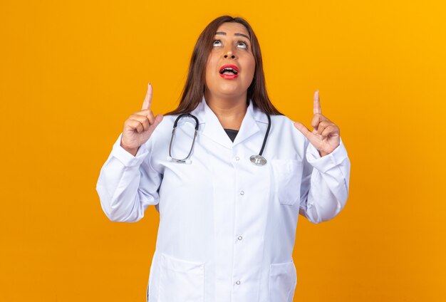 Femme médecin d'âge moyen en blouse blanche avec stéthoscope regardant surpris pointant avec l'index debout sur le mur orange