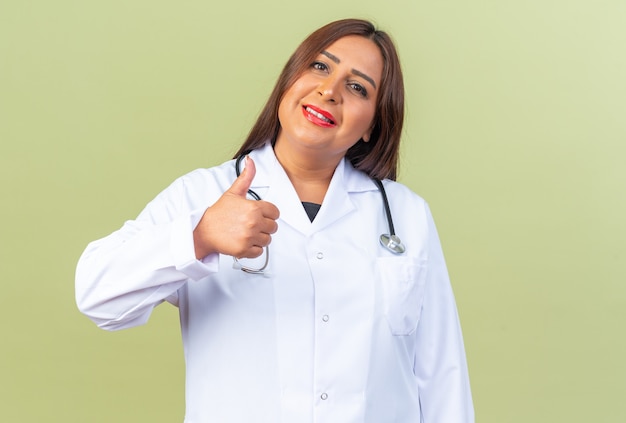 Femme médecin d'âge moyen en blouse blanche avec stéthoscope à sourire confiant montrant les pouces vers le haut