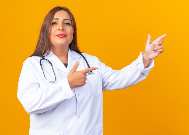 Femme médecin d'âge moyen en blouse blanche avec stéthoscope avec un sourire confiant pointant l'index sur le côté