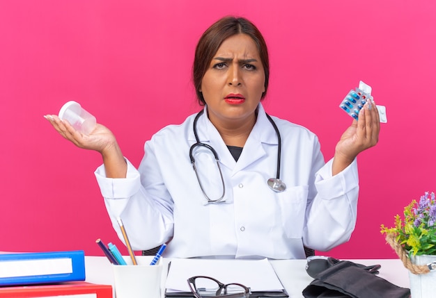 Femme médecin d'âge moyen en blouse blanche avec stéthoscope tenant différentes pilules et pot de test regardant devant confus assis à la table avec des dossiers de bureau sur un mur rose