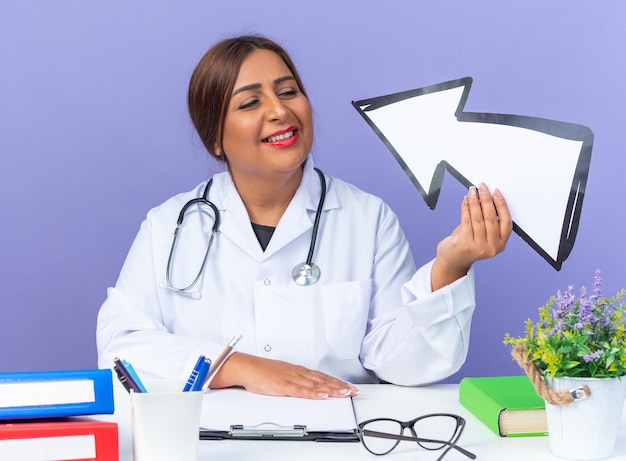 Femme médecin d'âge moyen en blouse blanche avec stéthoscope tenant du blanc en le regardant avec un sourire sur un visage intelligent assis à la table sur bleu