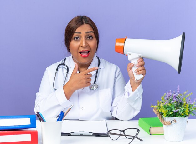 Femme médecin d'âge moyen en blouse blanche avec stéthoscope tenant un mégaphone pointant avec l'index sur elle heureuse et surprise assise à la table sur bleu