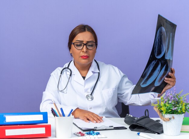 Femme médecin d'âge moyen en blouse blanche avec stéthoscope tenant une radiographie en regardant le presse-papiers sur la table avec un visage sérieux assis à la table sur un mur bleu