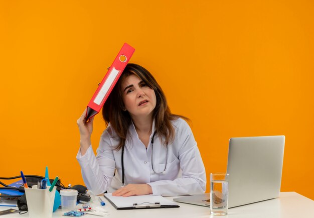 Femme médecin d'âge moyen concerné portant une robe médicale avec stéthoscope assis au bureau de travail sur un ordinateur portable avec des outils médicaux mettant le dossier sur la tête sur un mur orange isolé avec espace de copie